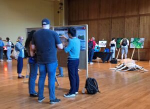 3 people stand in the foreground discussing a graduate student poster. In the background, people look at various art displays.