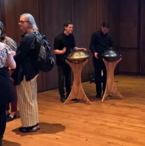 Robert and Chris play handpans. There is a group of people having a conversation in the foreground.