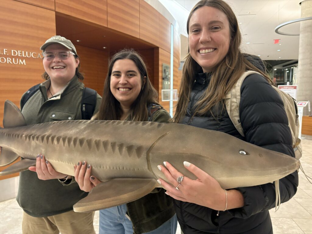 Three students holding a large foam model of a white sturgeon.